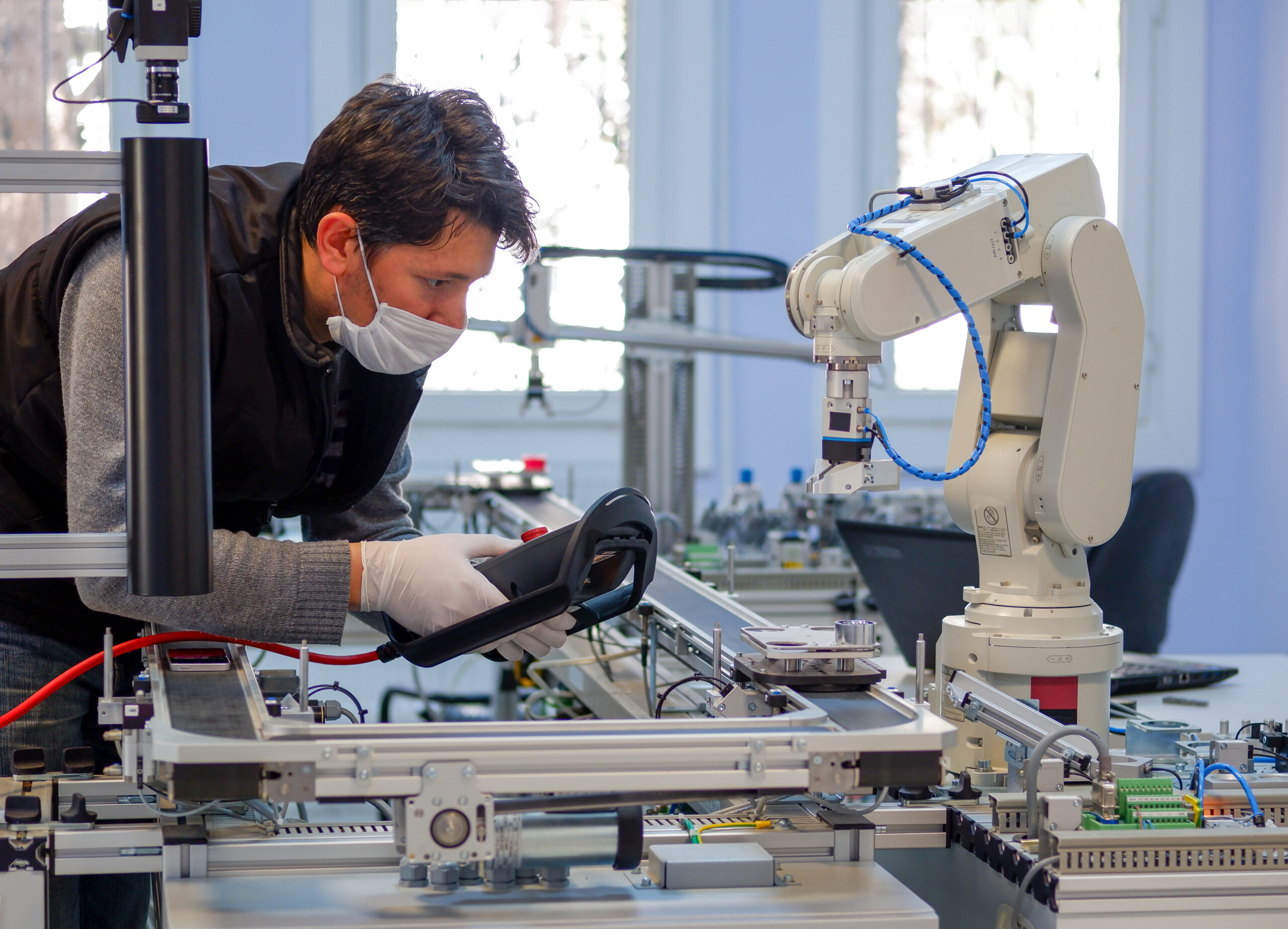 Man with surgical mask on face against COVID-19 ( Coronavirusis ) programming robot arm with control panel which is integrated on smart factory production line. industry 4.0 automation line.